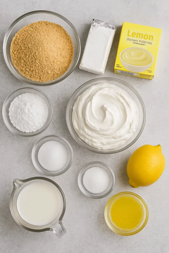 Ingredients for Lemon Lush Dessert arranged on a light gray surface, including graham cracker crumbs, cream cheese, lemon pudding mix, whipped topping, and fresh lemon.